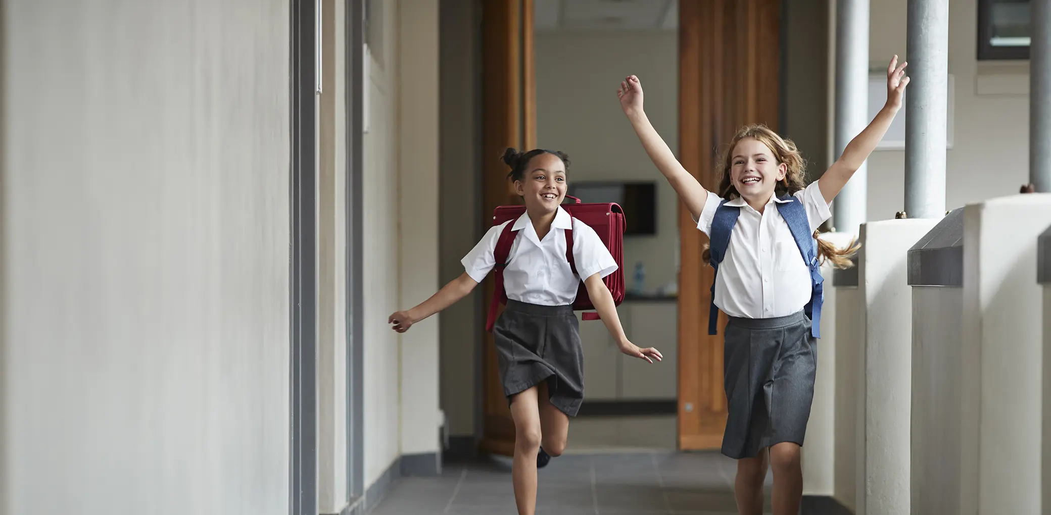 Two primary school girls skip together in a hallway.