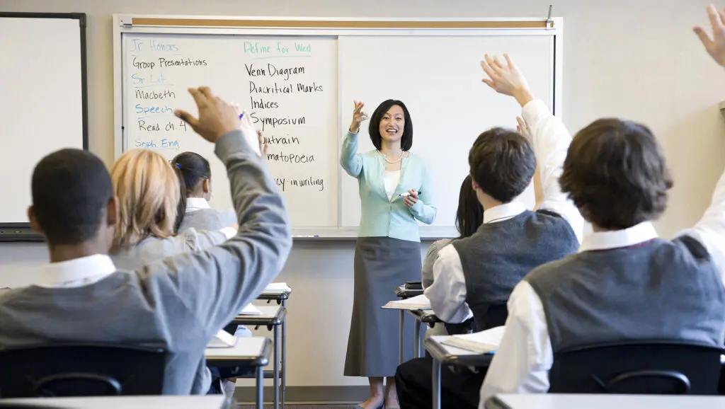 A high school teacher standing in front of a white board in a classroom calls on students with raised hands.
