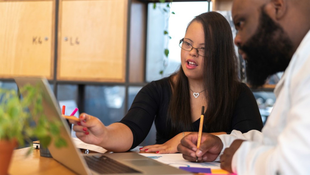 People with disabilities work together on a project, one person pointing to the screen on a laptop while the other writes on paper.