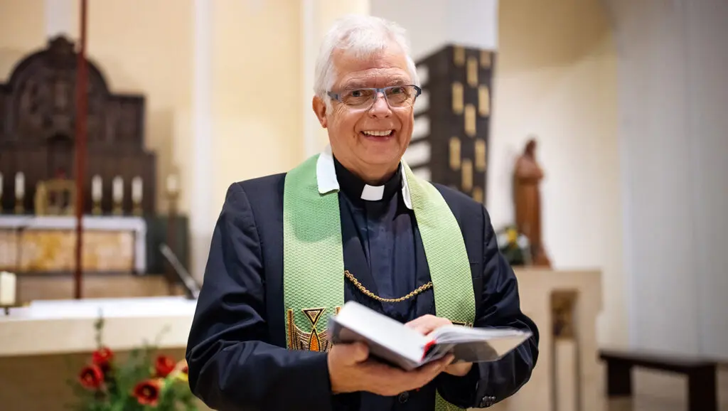 A Catholic priest stands in a church, smiling, as he holds open a bible.