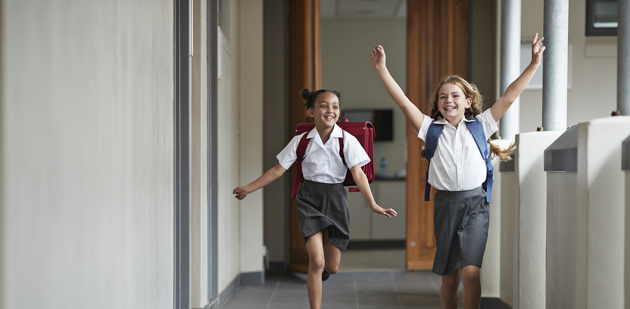 Two primary school girls skip together in a hallway.