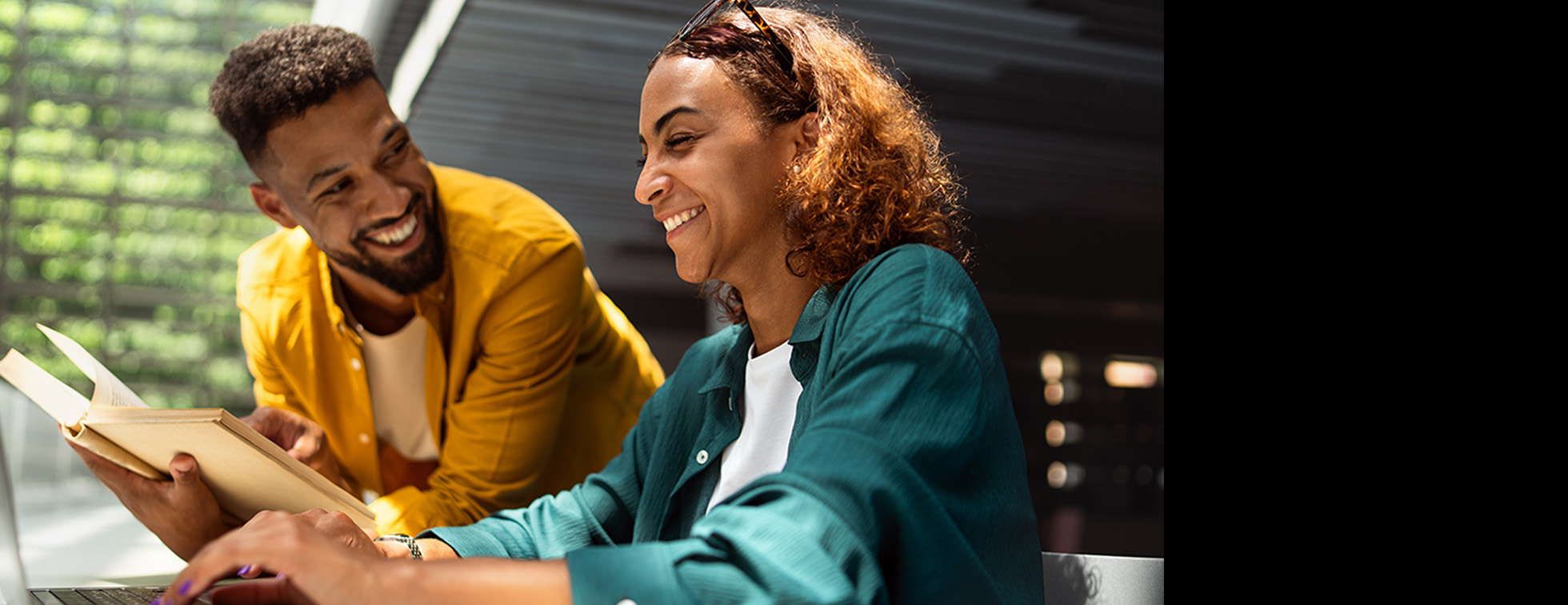 A picture of two people of color smiling together, one holding an open book and one typing at their laptop.