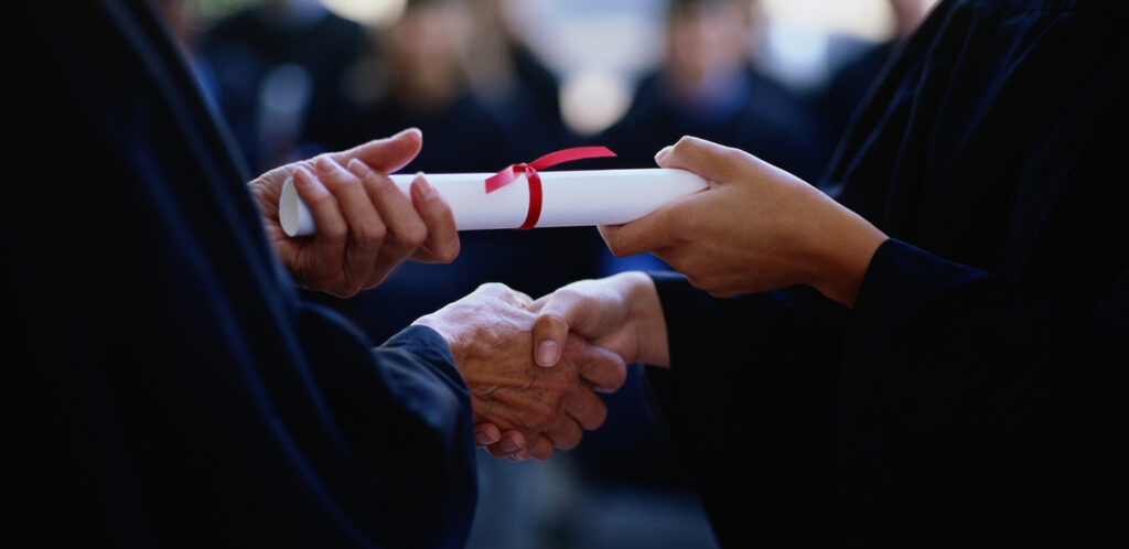A picture of a hand shake while passing a diploma, symbolizing leadership transition in higher education.